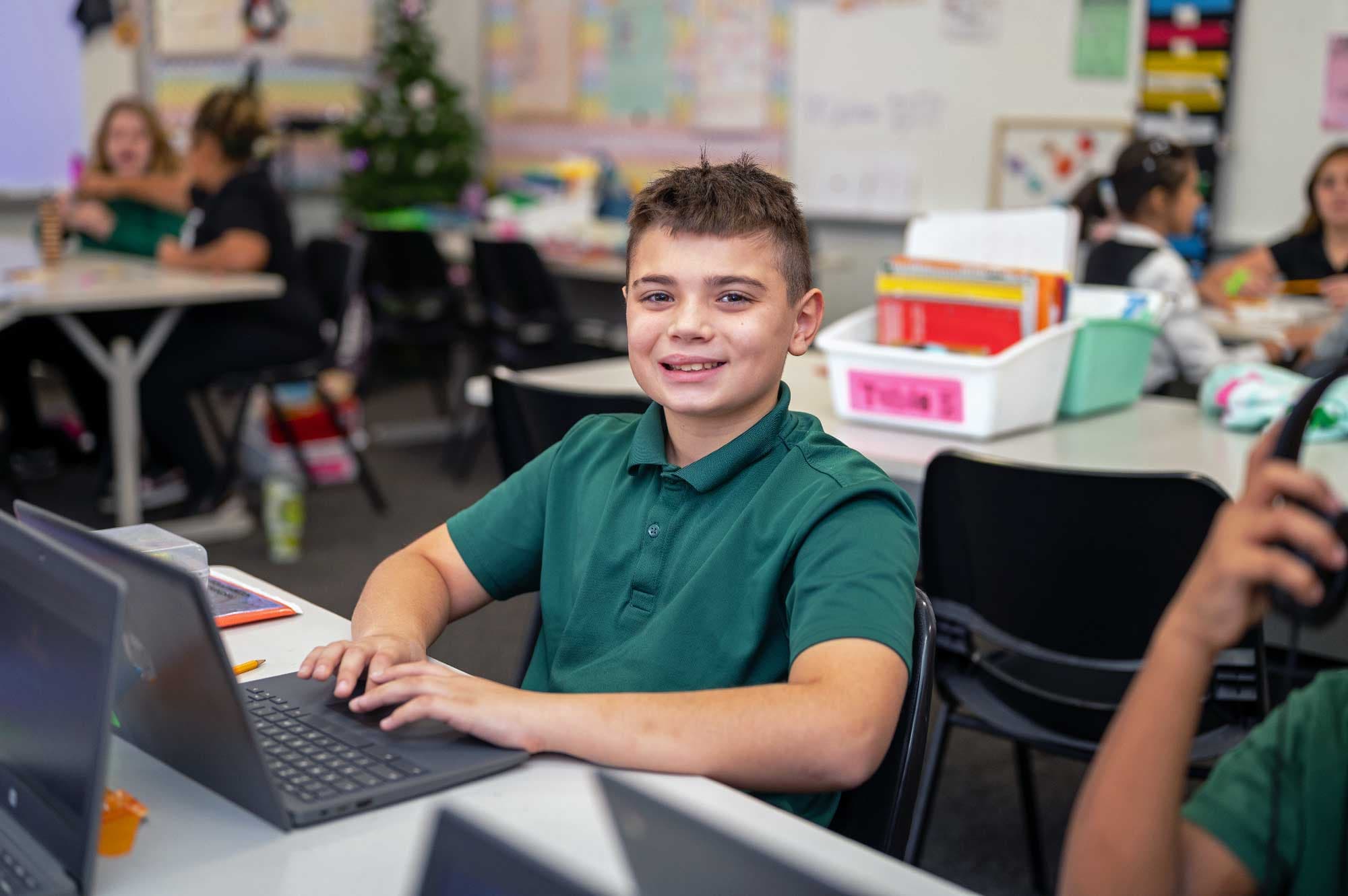 Student in a green school polo smiling while working on a laptop in a classroom.