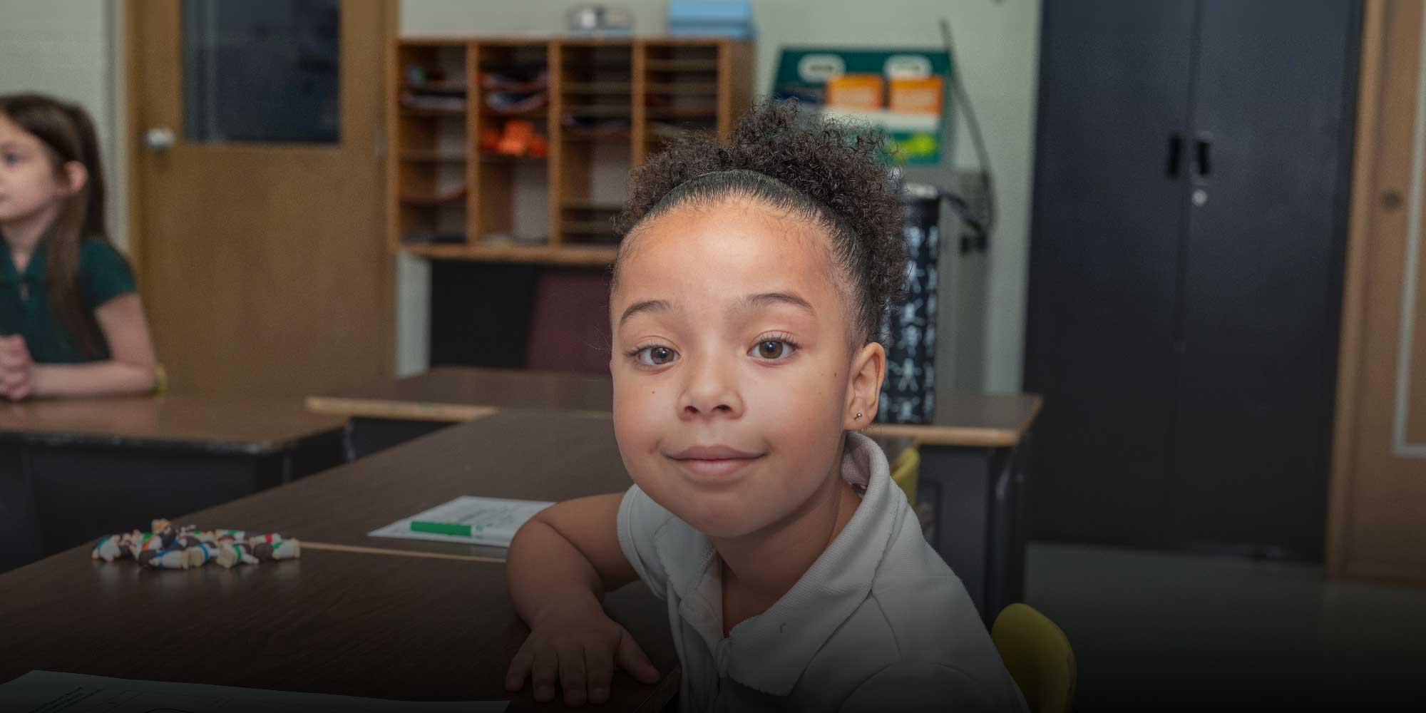 Young student smiling while seated at a classroom desk.